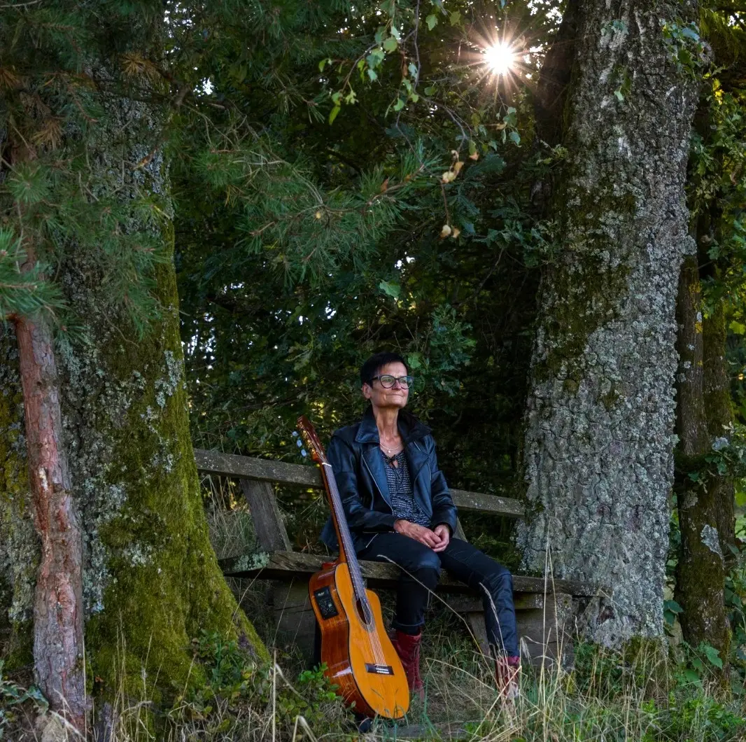 Singer Songwriterin Isolde, neben ihr angelehnt ihre braune Akustikgitarre, sitzt bei einem Sonnenuntergang zufrieden auf einer Holzbank zwischen zwei alten Birken und sieht in die Ferne. Zwischen dem Laub der Bäume blitzt die untergehende Sonne hervor.