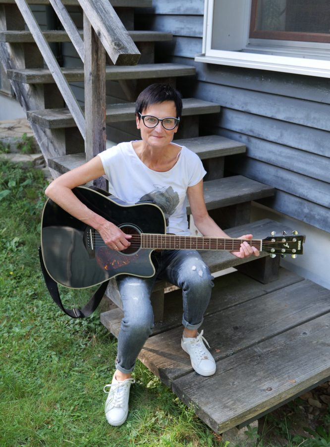 Singer Songwriterin Isolde sitzt mit schwarzer Akustikgitarre vor einem grauen Holzhaus auf einer Holztreppe