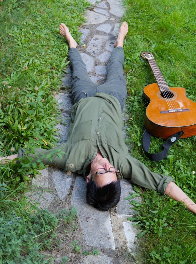 Singer Songwriterin Isolde liegt auf dem Rücken, die Arme und Beine von sich gestreckt, auf einem schmalen Gartenweg, rechts und links grüne Wiese, die Akustikgitarre liegt neben ihr im Gras und sie sieht in den Himmel