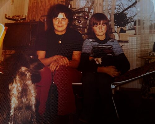 Isolde als etwa 11-jähriges Mädchen sitzt mit ihrer Mutter im Wohnzimmer auf eine Liege, mit dabei sind ihr Hund, ein schwarz-weißer Cockerspaniel und ihre Katze